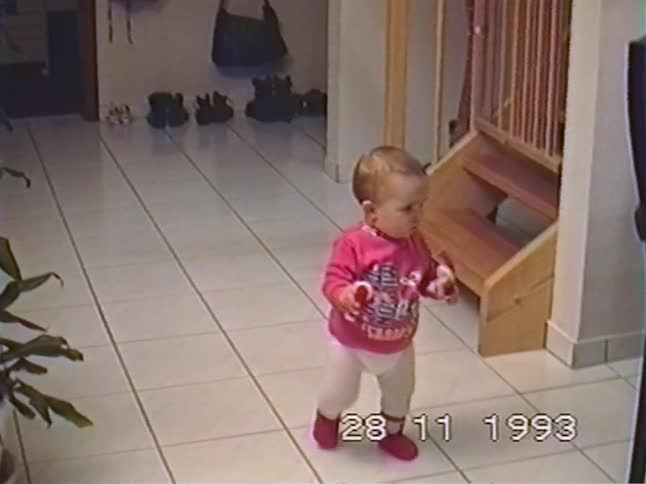 A vintage home video from 1993 captures the precious moment a baby girl takes her first wobbly steps across the tiled floor, holding a toy in each hand
