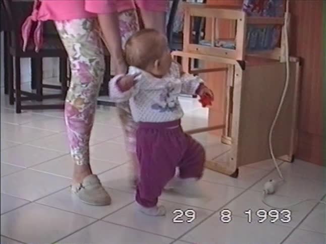 A proud mother helps her happy baby take its first steps across the living room floor in a vintage 1993 home movie