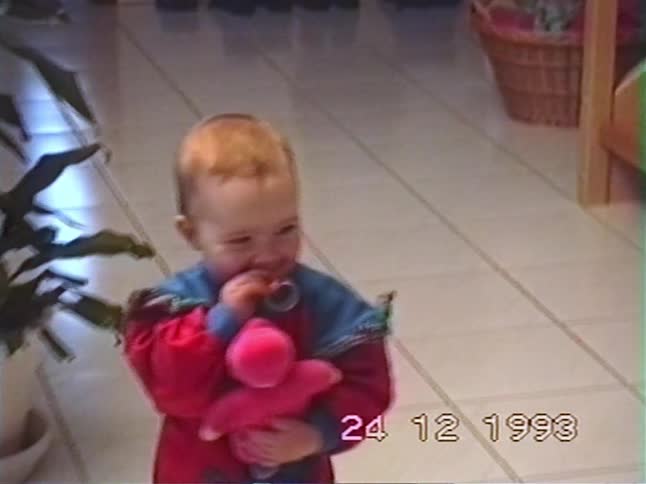 An adorable toddler with a pacifier holds a pink stuffed toy and smiles for the camera in a nostalgic home video from 1993