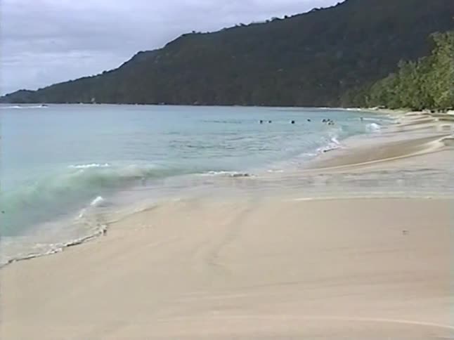 Serene vintage footage of gentle waves lapping a sandy tropical shore with a lush green hillside in the background