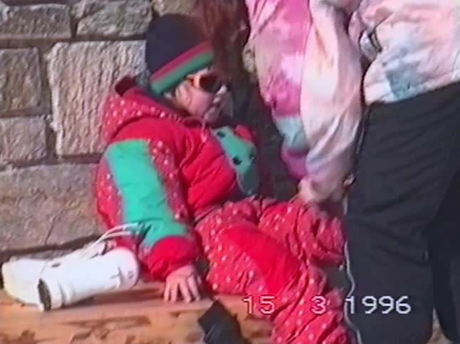 A cute little girl in a bright red snowsuit and sunglasses smiles while sitting on a bench at a ski resort