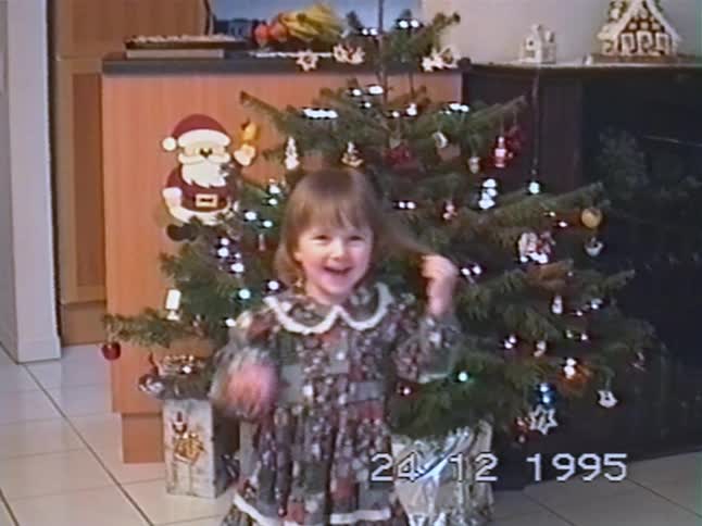 A cute little girl in a festive dress smiles and plays with her hair in front of a decorated Christmas tree in a vintage 1995 home video