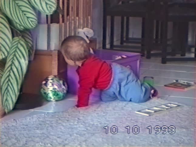 A cute baby in a red shirt plays with toys on the floor before crawling and trying to stand up in a living room in 1993