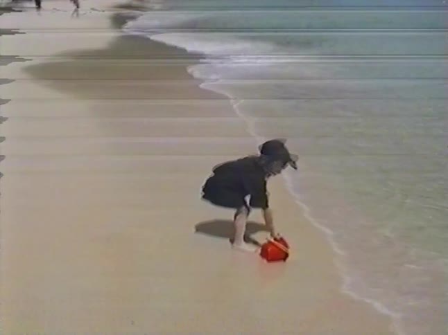 Vintage footage shows a little girl in a baseball cap filling her red bucket with water at the edge of a tropical sea