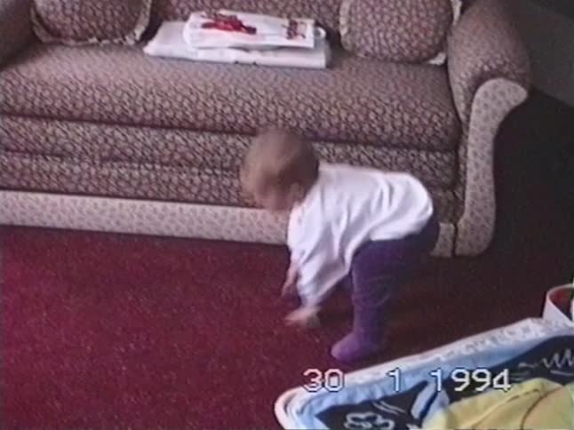 Vintage 1994 home video footage captures a cute baby learning to stand up for the first time on a red carpet in the living room