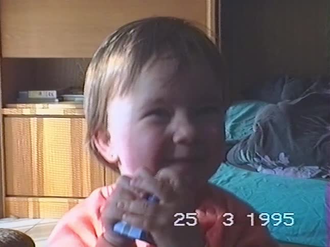 A close-up of a cute, smiling toddler with rosy cheeks in an orange sweater from a 1995 home video