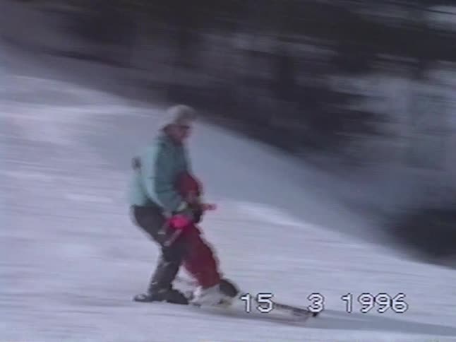 A man patiently teaches a toddler how to ski by holding them as they glide down a snowy hill in a vintage 1996 home video