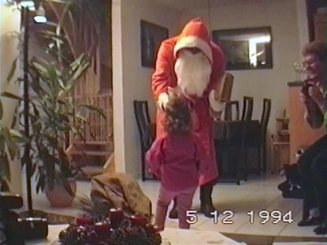A person dressed as Saint Nicholas reads from a book to a little girl during a family holiday celebration in a vintage 1994 home movie