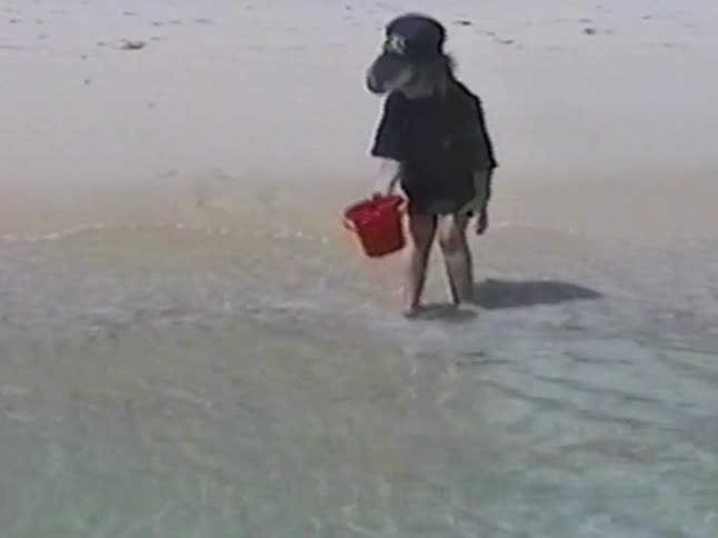 Vintage footage shows a young girl with a red bucket letting the ocean waves wash over her feet on a sandy beach
