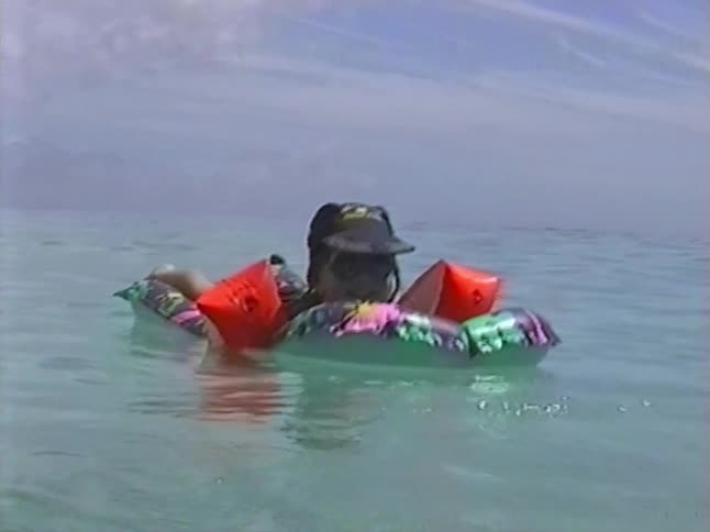 Vintage home movie footage from the 1990s shows a young girl with arm floaties relaxing on an inflatable raft in the calm ocean