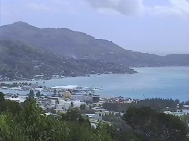 A vintage VHS pan shot reveals a tropical island city with a bustling harbor at the base of lush green mountains