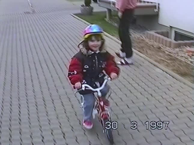A happy young girl wearing a colorful helmet rides her bike toward the camera on a paved driveway in this 1997 home movie