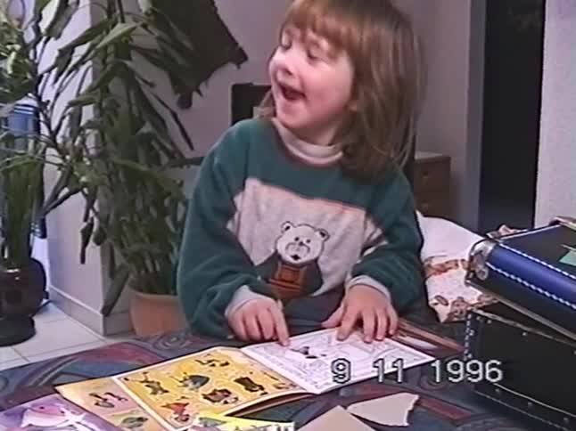 An adorable little girl in a teddy bear sweater smiles while playing with a sticker book in a 1996 home movie