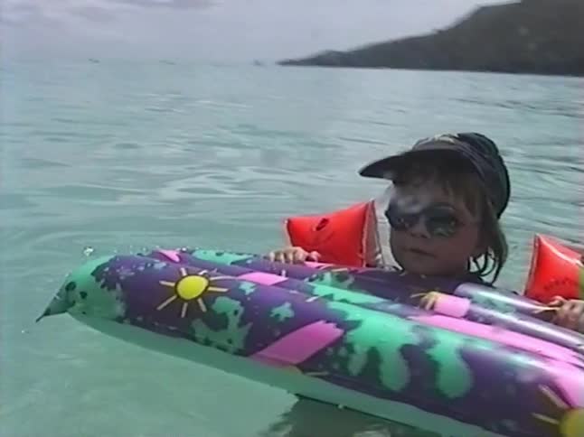 Vintage VHS footage captures a joyful young girl with sunglasses and arm floaties floating on a raft in a tropical ocean