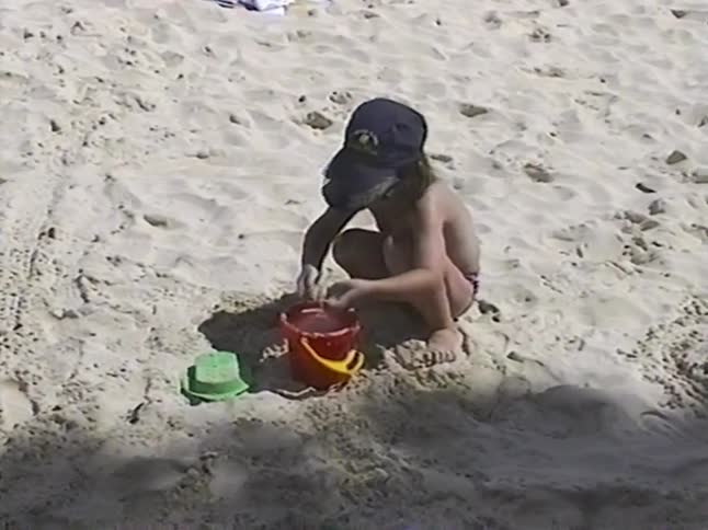 Vintage home movie footage captures a little girl in a hat and sunglasses playing with a bucket in the sand before smiling at the camera