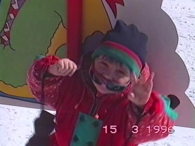 A young child in a colorful 90s snowsuit happily dances and plays with their sunglasses on a snowy ski slope