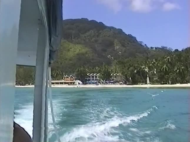 A vintage VHS view from a departing boat captures the wake trailing in the turquoise water towards a scenic tropical island resort