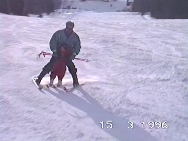 A father teaches his young child how to ski down a snowy slope in this authentic home video from 1996
