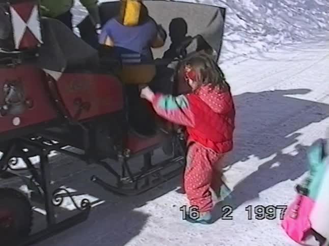Young children in colorful winter gear climb into an old-fashioned sleigh in the snow, from a 1997 home video