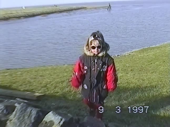 A young girl in a cool winter coat and sunglasses poses by the water's edge on a windy day in 1997
