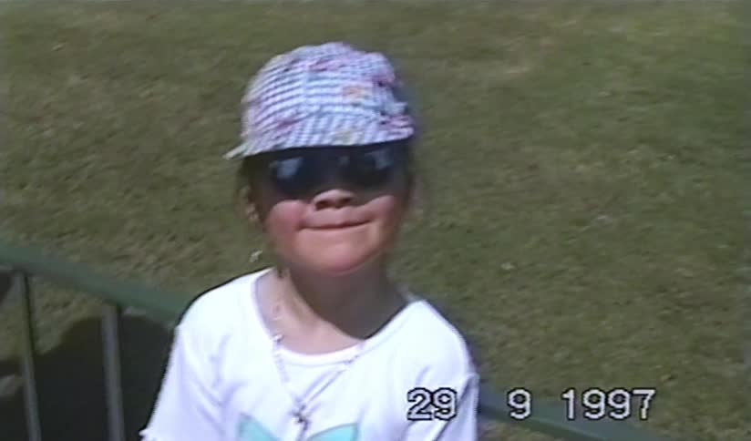 A cute little girl in a hat and sunglasses gives a cheesy grin for the camera in this nostalgic 1990s home video