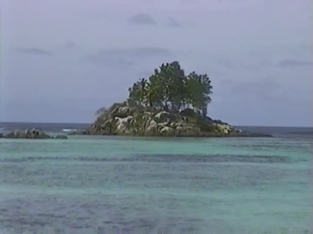 A slow pan across a calm tropical bay reveals a small rocky island with palm trees, captured on vintage VHS tape
