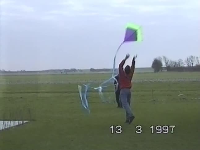 Archival footage from 1997 shows two men launching a colorful kite in slow motion in a large grassy field