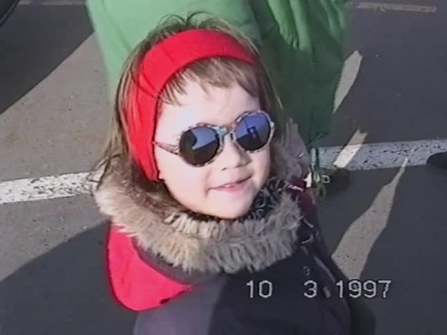 A happy little girl in a winter coat and sunglasses smiles for the camera in this authentic home movie from 1997