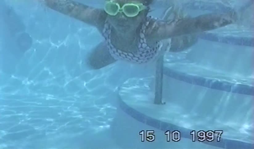 A young girl with goggles swims underwater towards the camera in a swimming pool on vintage home video from 1997