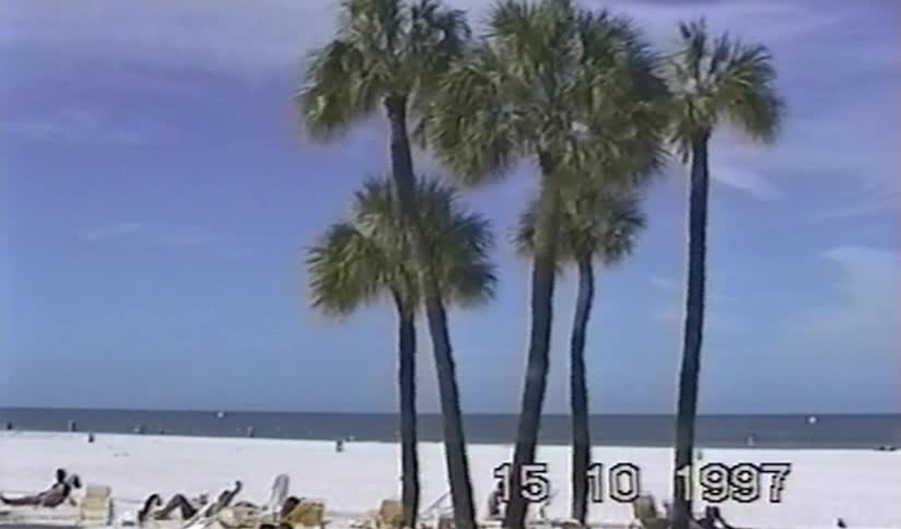 A vintage VHS shot pans across a sunny white sand beach with palm trees and sunbathers by the ocean in 1997