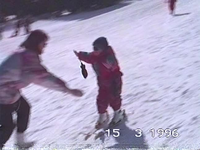 A mother catches and hugs her young child as they learn to ski down a snowy hill in this nostalgic home video from 1996