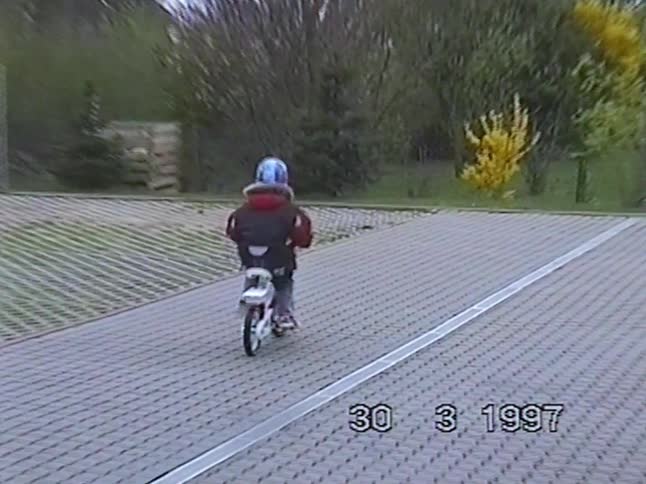 Vintage 1997 home video shows a mother pushing her daughter on a bike, who then rides independently through a paved lot