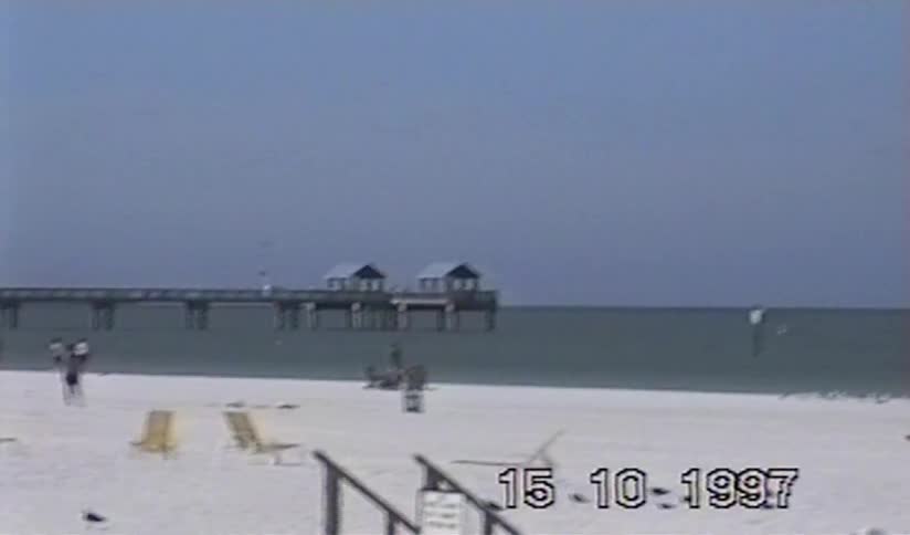 Nostalgic VHS footage from 1997 pans across a white sand beach with a long pier stretching into the ocean
