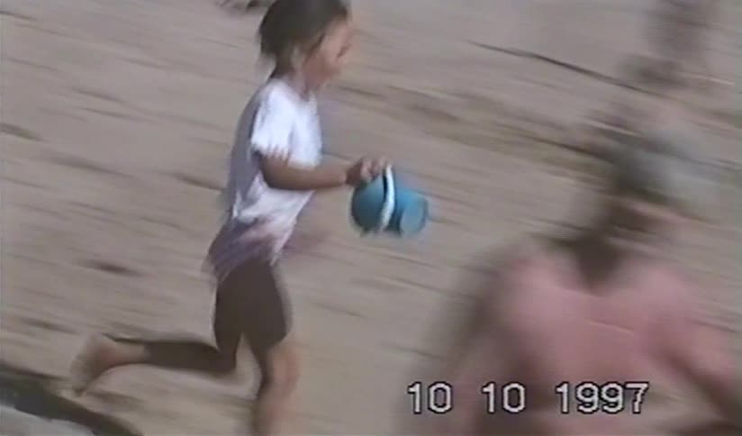 A young girl with a bucket runs happily along a sandy beach in a nostalgic home video from 1997