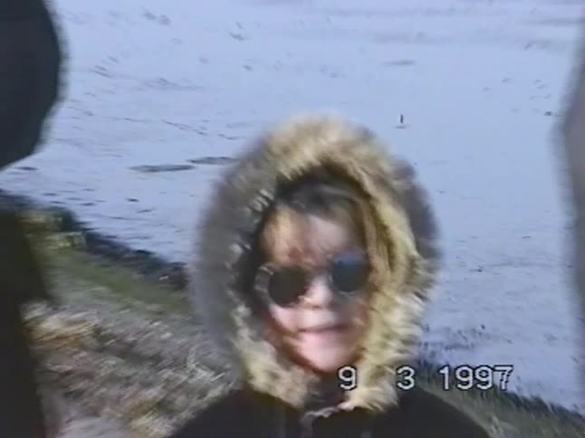 A vintage 1997 clip shows a happy young girl in a winter coat with a fur hood and sunglasses posing by the sea