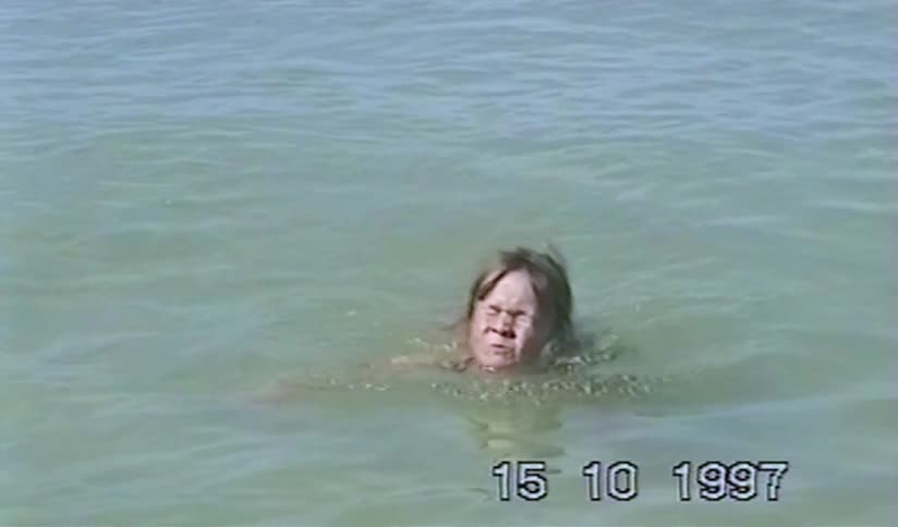Vintage home video from 1997 shows a young girl with braided hair splashing and swimming in the calm ocean