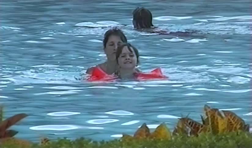 A mother helps her smiling young daughter with arm floaties swim in a pool in this nostalgic home video footage