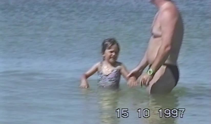 A young girl with pigtails laughs and plays in the ocean with her father in a nostalgic home video from 1997