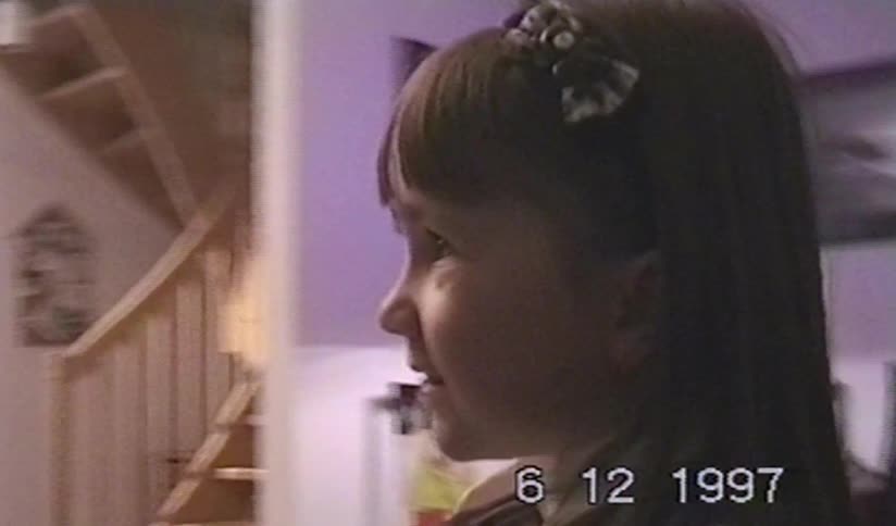 A nostalgic VHS clip from 1997 captures a young girl with a bow in her hair smiling inside her home