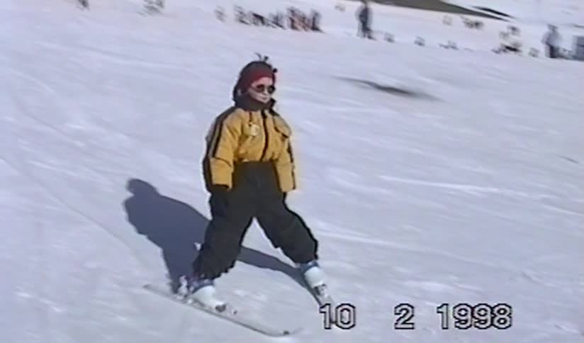 A vintage home video from 1998 captures a young girl skiing down a snowy mountain slope on a sunny day