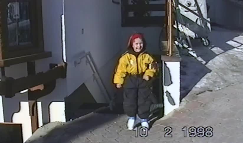 A happy little girl dressed in a yellow ski suit and red hat smiles for the camera on a sunny winter day in 1998