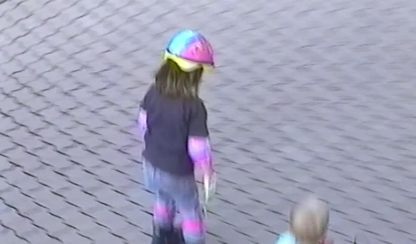 A high-angle vintage home video captures a young girl in a colorful helmet and pads learning to rollerblade on a paved driveway