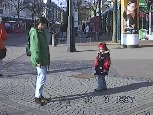 A mother and her child stand on a sunny cobblestone street in a European city in 1997