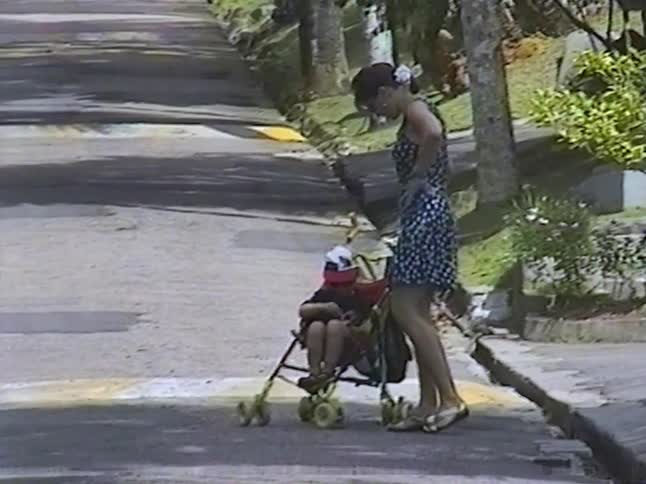 A mother in a floral dress stands with her son in a stroller on a tree-lined street in this vintage home video