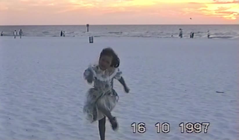 A little girl in a dress runs joyfully in slow motion towards the camera on a windy beach at sunset in this nostalgic 1997 home video