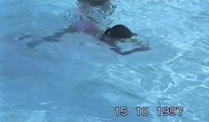 Vintage 1997 home movie shows a father teaching his young daughter with green goggles how to swim and go underwater in a pool