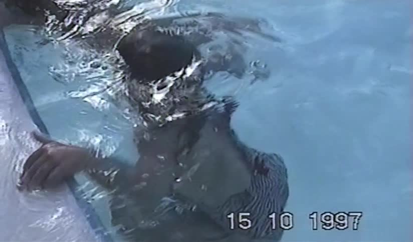 A vintage home movie from 1997 shows two children learning to snorkel and swim in a pool on a sunny day