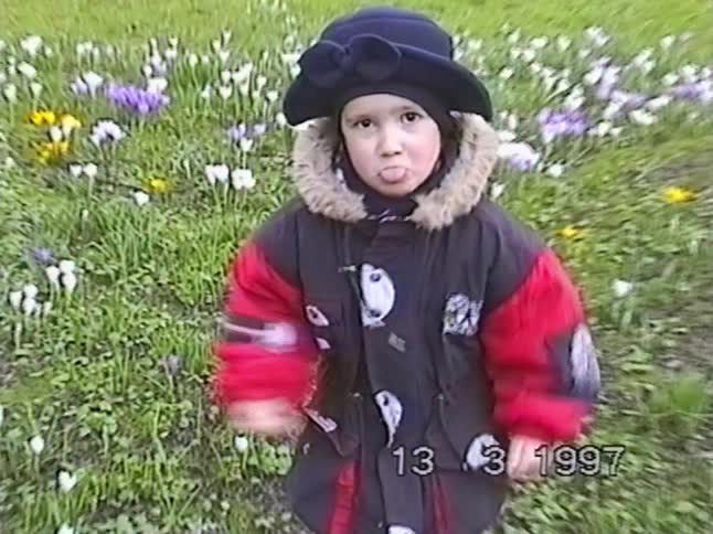 Vintage 1997 VHS footage of a cute little girl in a winter coat and hat playfully sticking her tongue out while standing in a field of spring crocus flowers