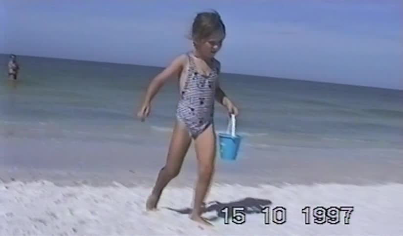 Archival footage from 1997 shows a young girl in a swimsuit walking out of the ocean and onto a sandy beach with a small bucket