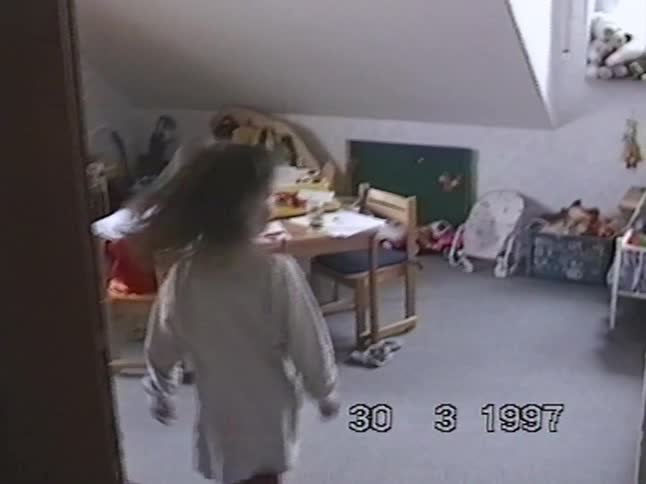 A little girl crawls across the carpeted floor of her messy playroom in a home video from 1997